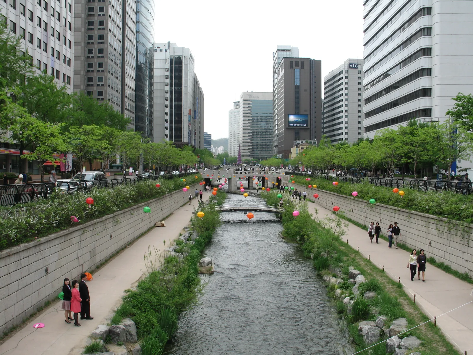 Colourful lanterns strung over Cheonggyecheon, with people walking beside the stream and Seoul buildings in the background